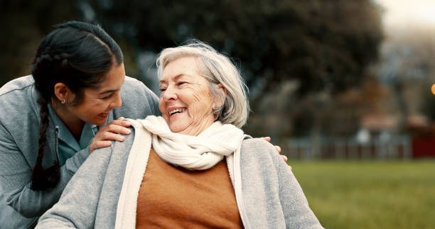 Young woman smiles at an elderly woman in a wheelchair, touching her shoulder outdoors.