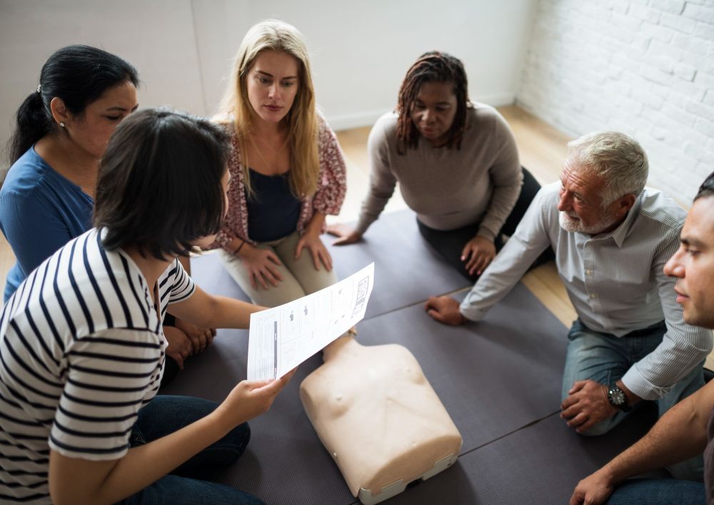 Group of people in a CPR training class, looking at a training dummy and documents on the floor.