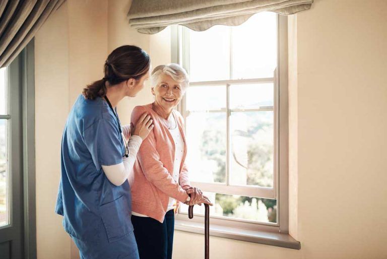 Nurse assisting an elderly person with a cane near a window. Sunlight illuminates them; both smile.