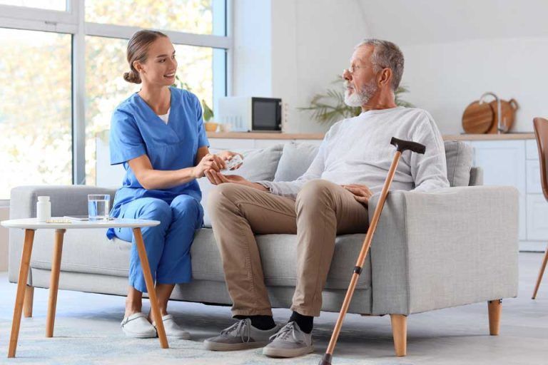 Caregiver holding hand of person sitting on a couch, indoors. A cane rests nearby.
