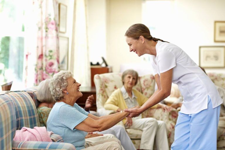 Nurse smiling and holding hands with a seated woman, in a senior living room with other residents.