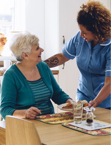 Caregiver serves a meal to an older adult at a table in a home setting.