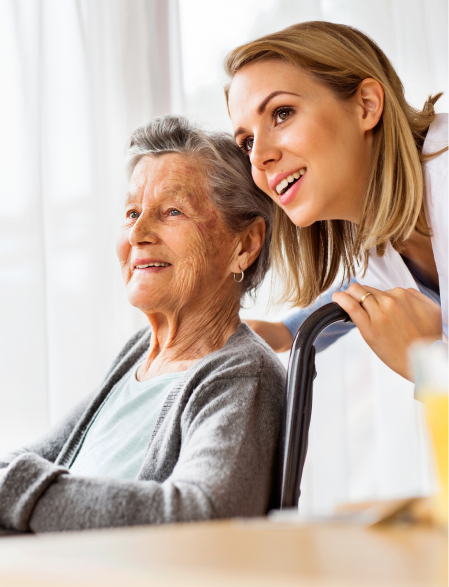 Woman in wheelchair with caregiver, both smiling, looking out a window.