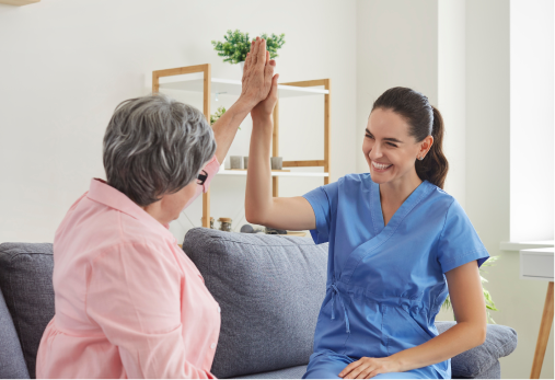 Woman in blue scrubs high-fives elderly woman on a couch; both smiling in a living room.