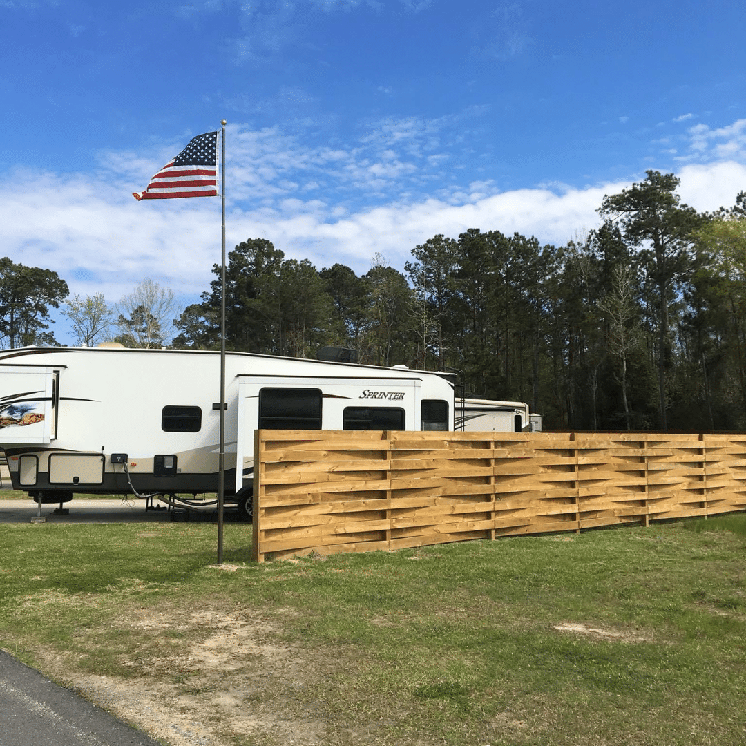 A rv is parked in a grassy field next to a wooden fence and an american flag.