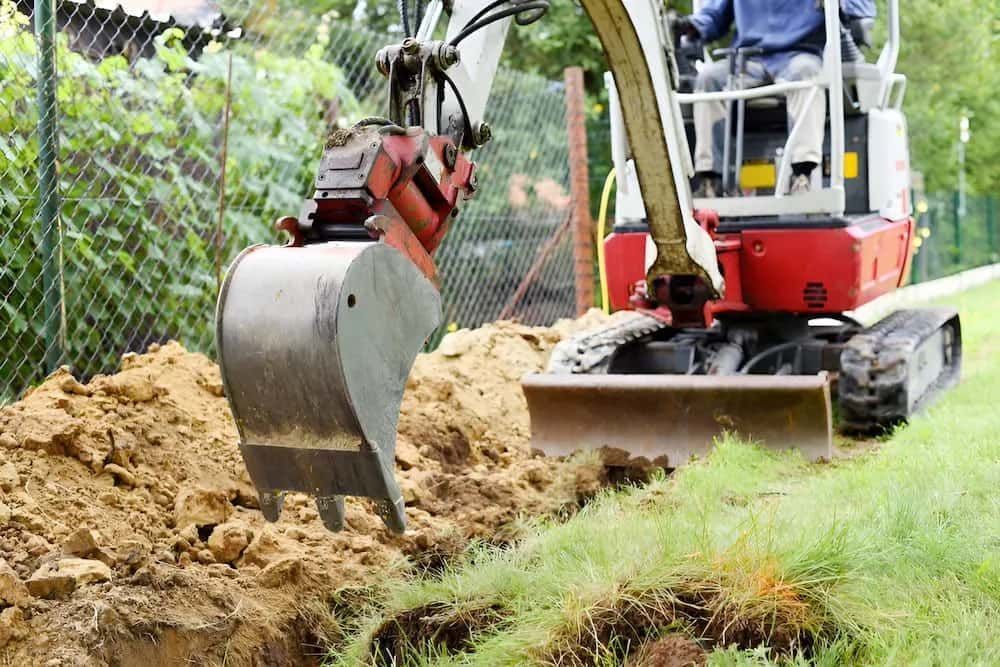 A Man Is Driving a Small Excavator Digging a Hole in The Ground — Jeff Thompson Excavator Hire Pty Ltd In Pink Lily, QLD
