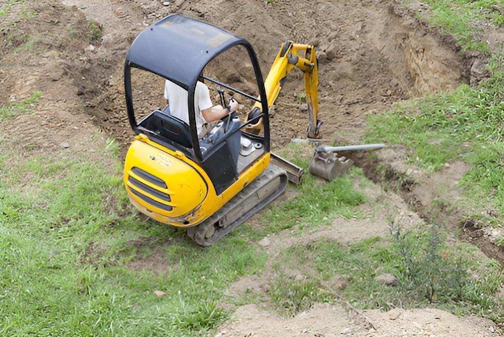 A Man Is Driving A Small Yellow Excavator In A Grassy Field — Jeff Thompson Excavator Hire Pty Ltd In Pink Lily, QLD