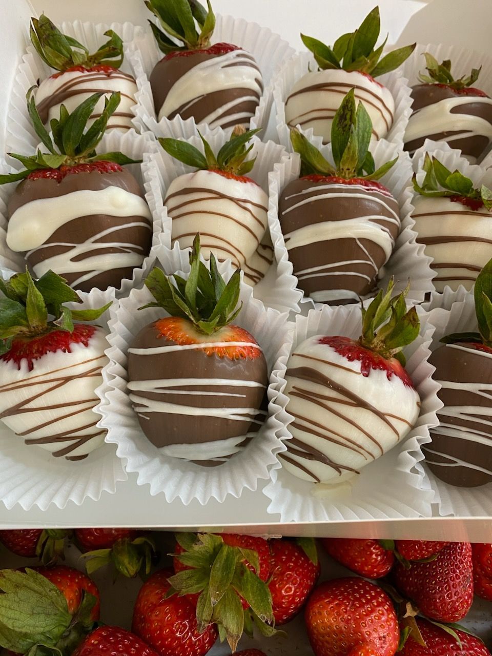 Chocolate-covered strawberries in a white box, decorated with white and dark chocolate drizzles, next to fresh strawberries.