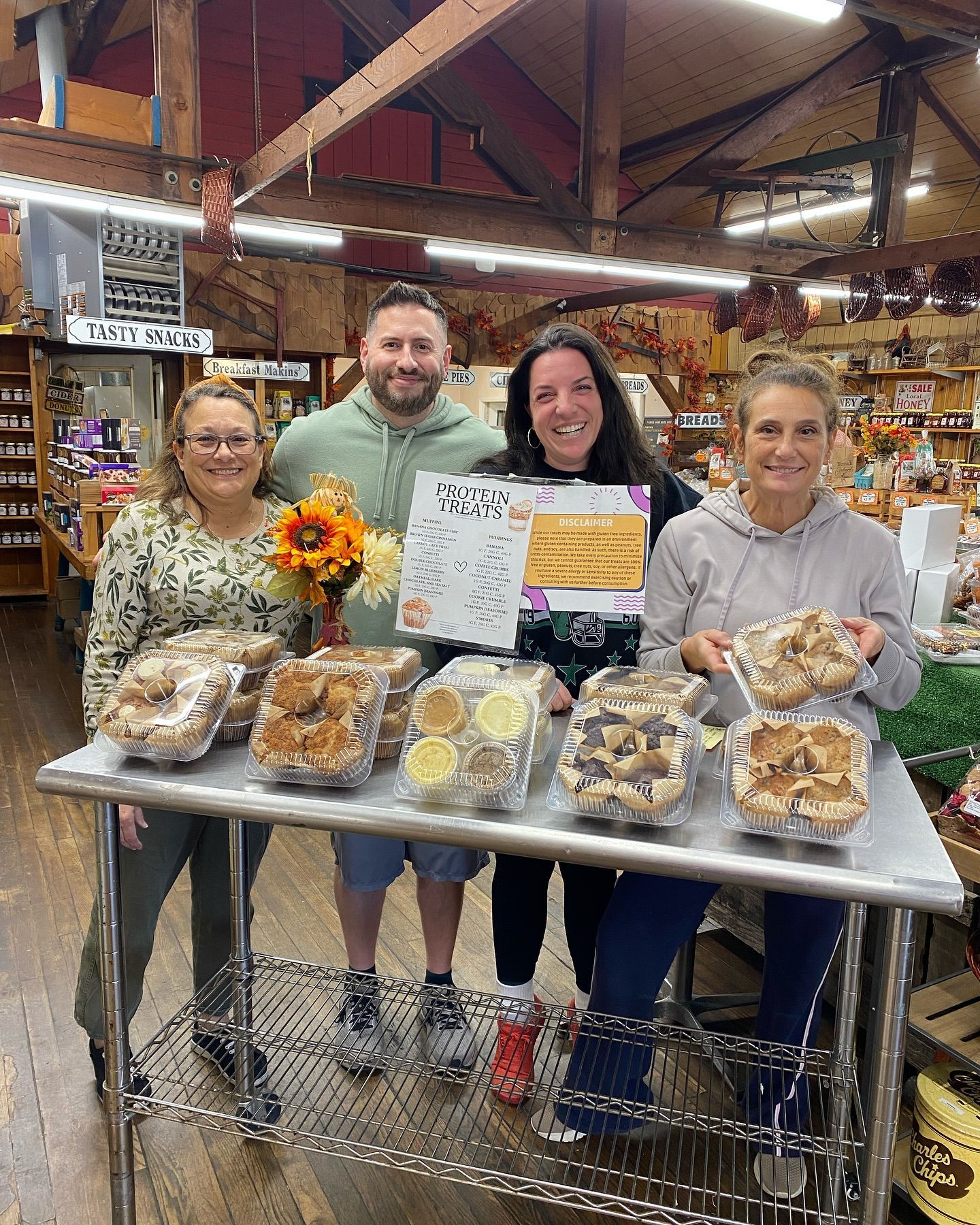 Four people in a store, holding and displaying various packaged food products on a stainless steel table.
