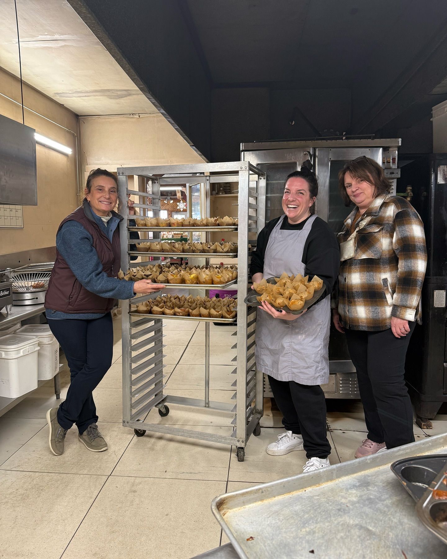 Three women smiling in a kitchen holding trays of muffins and showing them off.