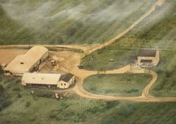 Aerial view of a farm with several buildings, a driveway, and surrounding green fields.