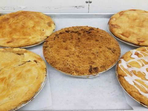 Five freshly baked pies on a metal tray; including a crumb-topped pie and glazed pie.