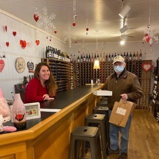 Woman and man in a wine shop. The woman is behind the counter. Man is holding a bag and a box.