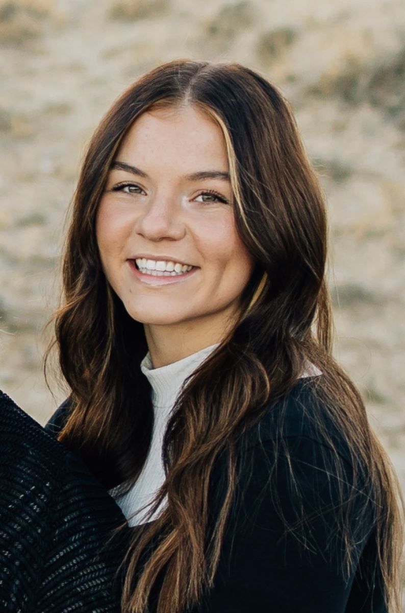A woman with long brown hair is smiling and wearing a black sweater.