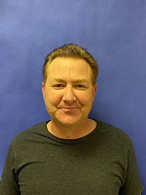 A man in a grey shirt is smiling in front of a blue wall.