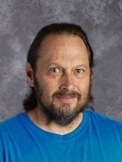 A man with a beard and long hair is wearing a blue shirt.
