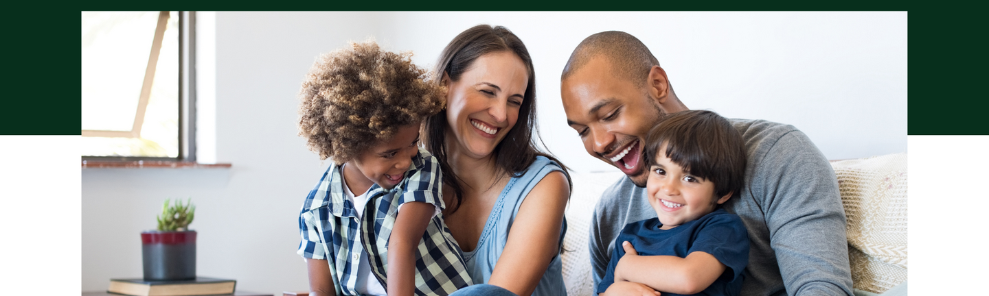 Family of four sitting on a couch, smiling and hugging in a bright living room.