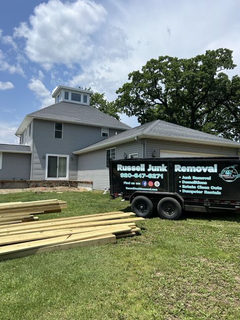 A Russell Junk Removal trailer parked in front of a grey two-story house under renovation with wood planks in the yard.