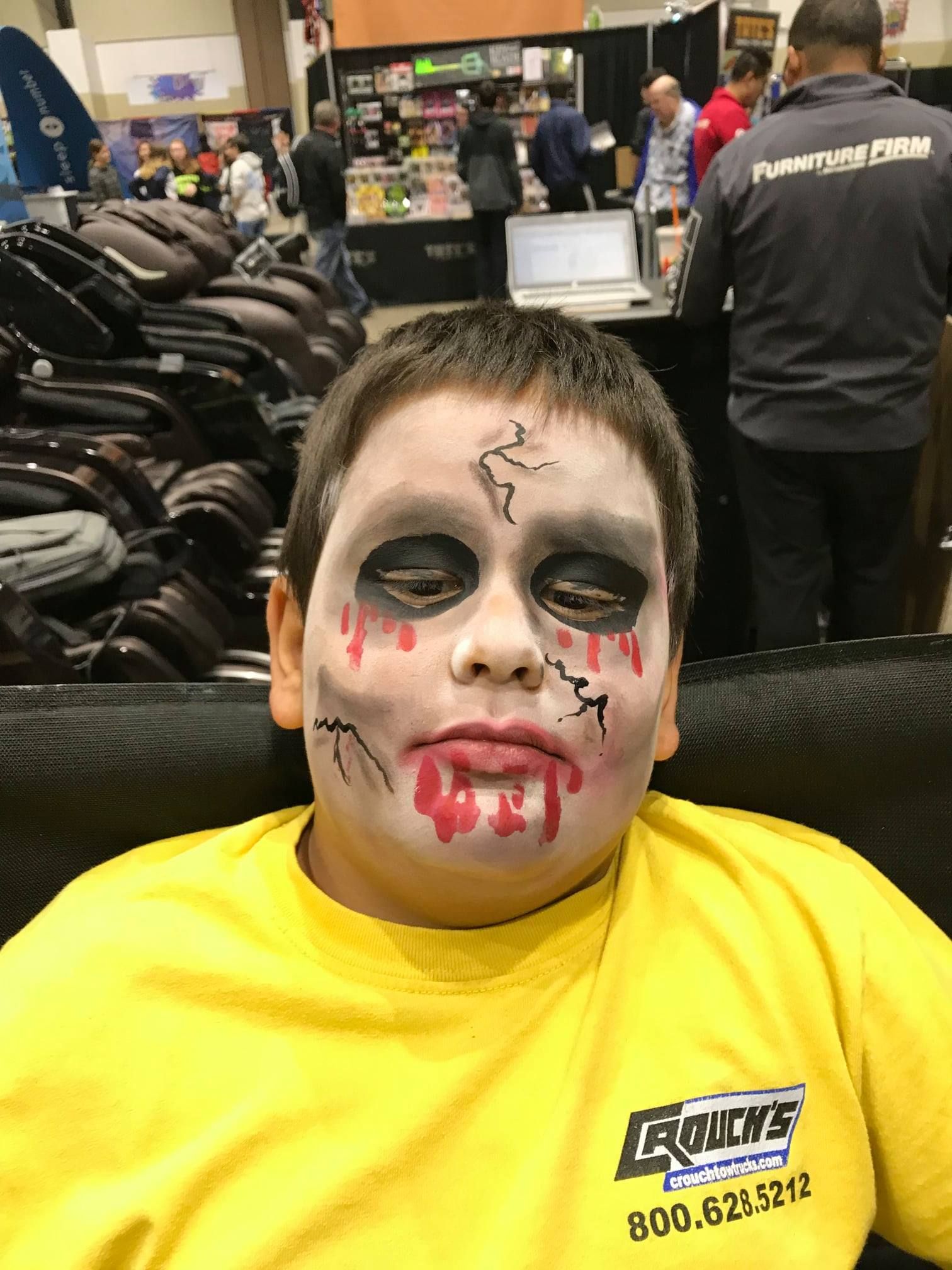 A person with zombie-style face paint and a yellow shirt sits in a room with rows of massage chairs in the background.