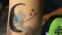 A sparkly temporary tattoo on skin depicting a silver fairy sitting on a black crescent moon with three bronze stars.