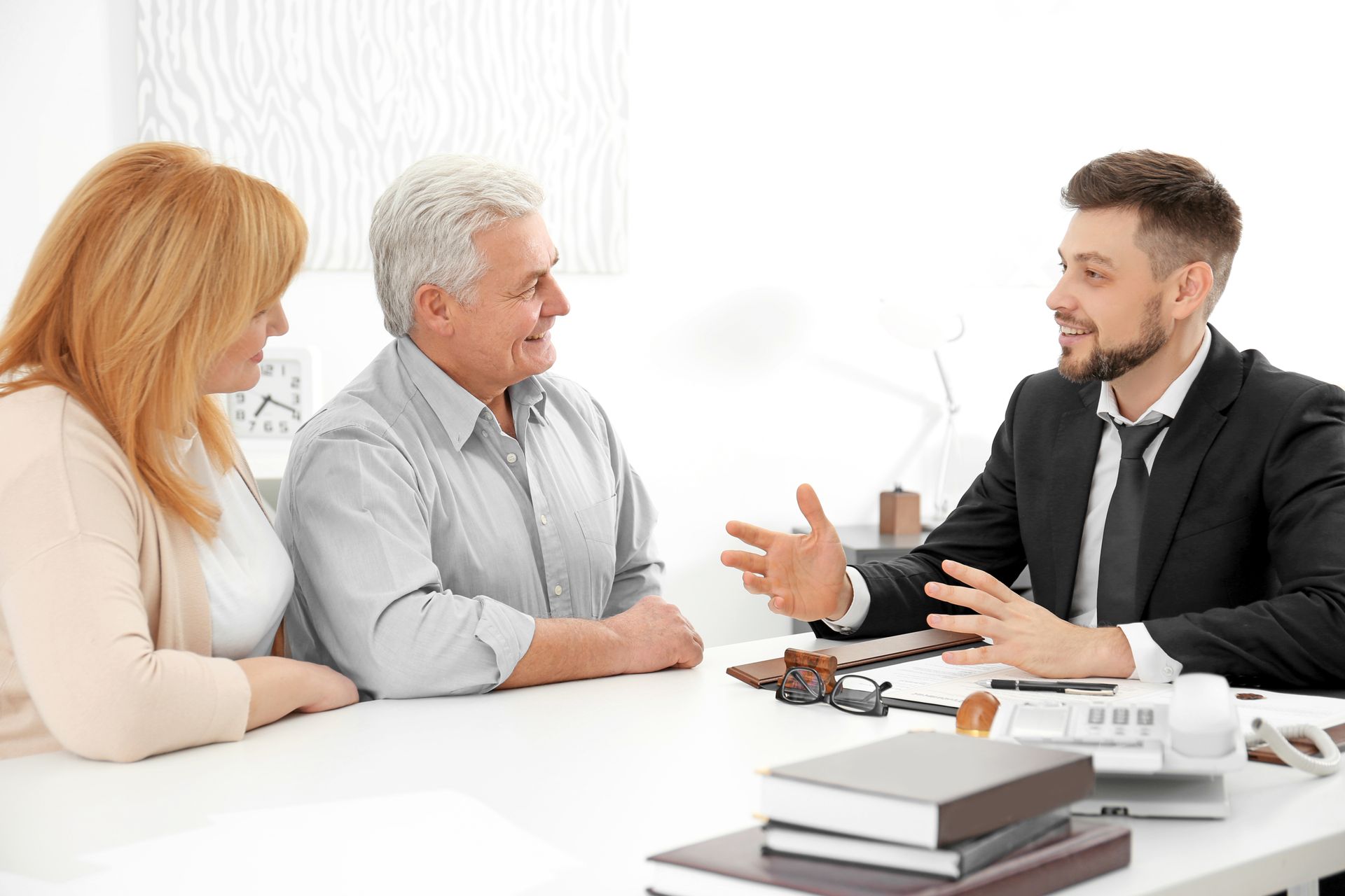 Three people in a bright office meeting, talking across a white table with notebooks and papers.