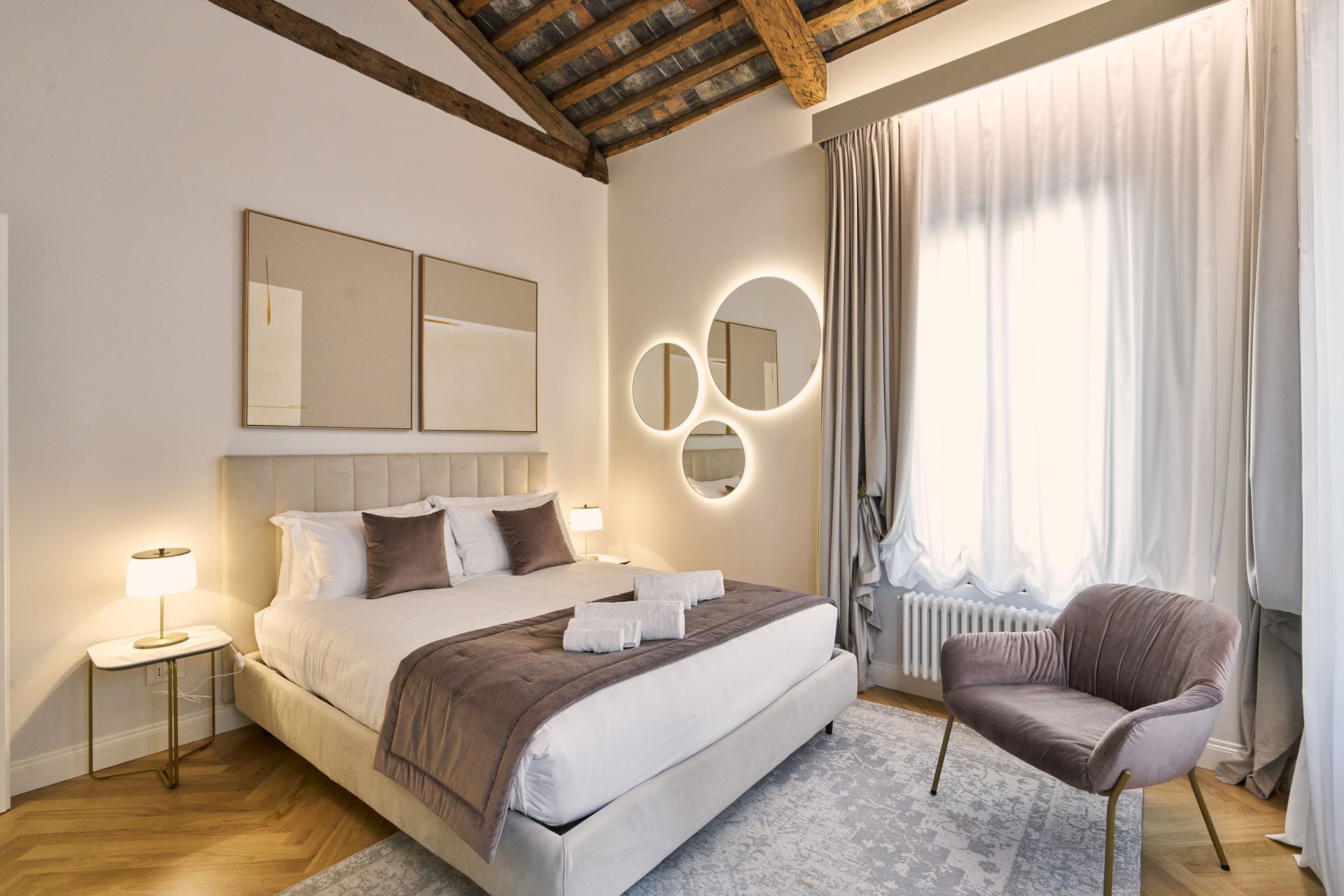 Modern bedroom with a bed, bedside lamp, round mirrors, and a chair by a large curtained window.