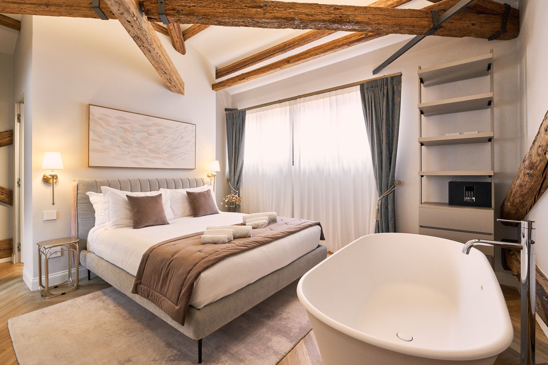 Cozy bedroom with exposed beams, a white bed, sheer curtains, and a freestanding bathtub in the foreground