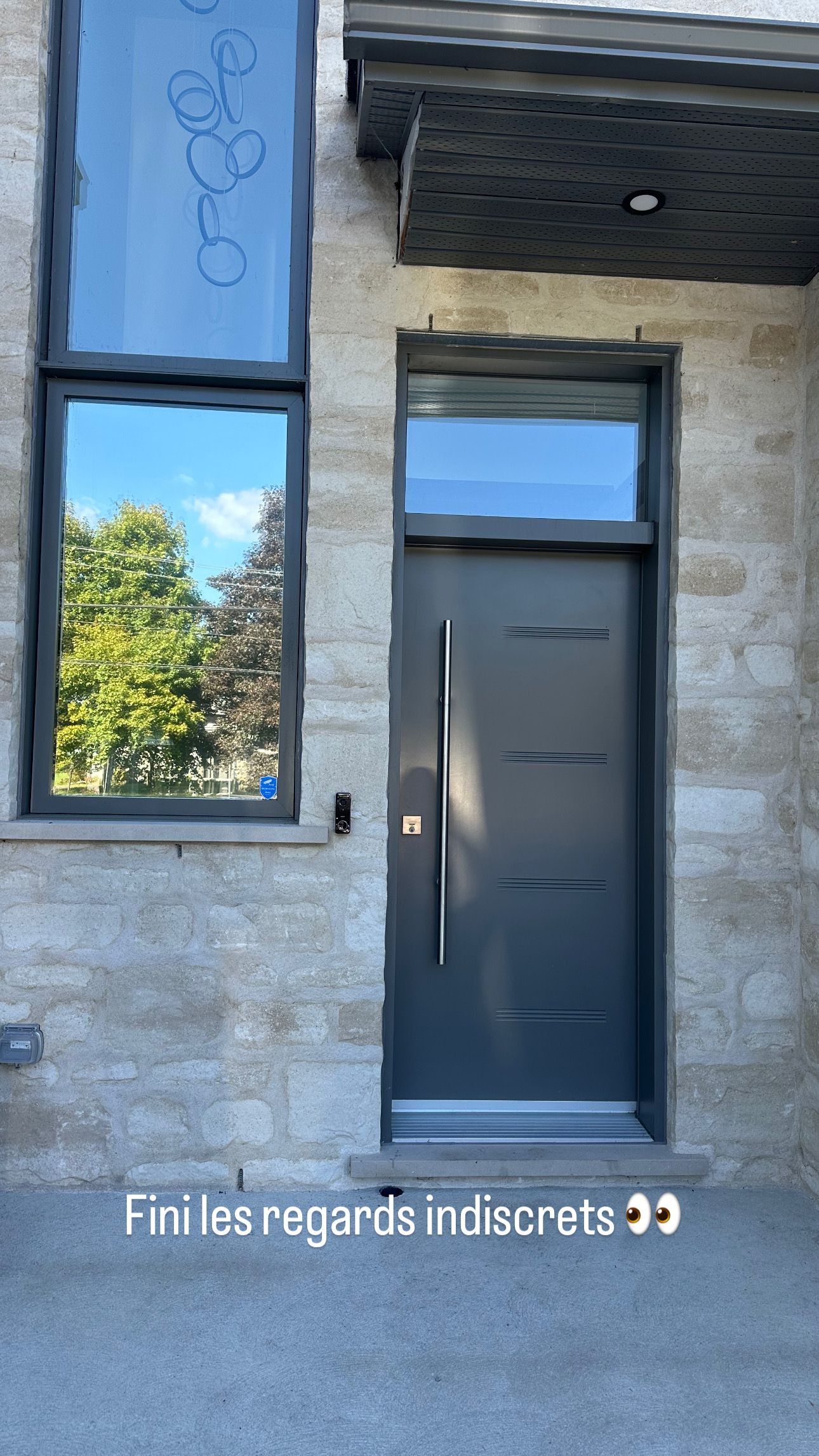 Porte d'entrée grise avec imposte et fenêtre latérales ; façade en pierre reflétant les arbres.