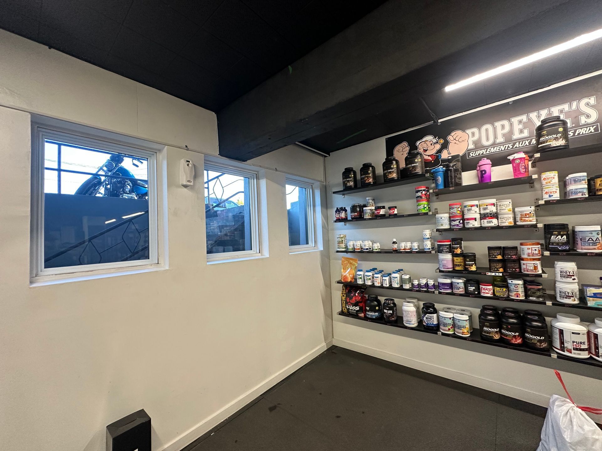 Intérieur d'un magasin de compléments alimentaires Popeye's avec des étagères de produits, trois petites fenêtres et un plafond sombre.