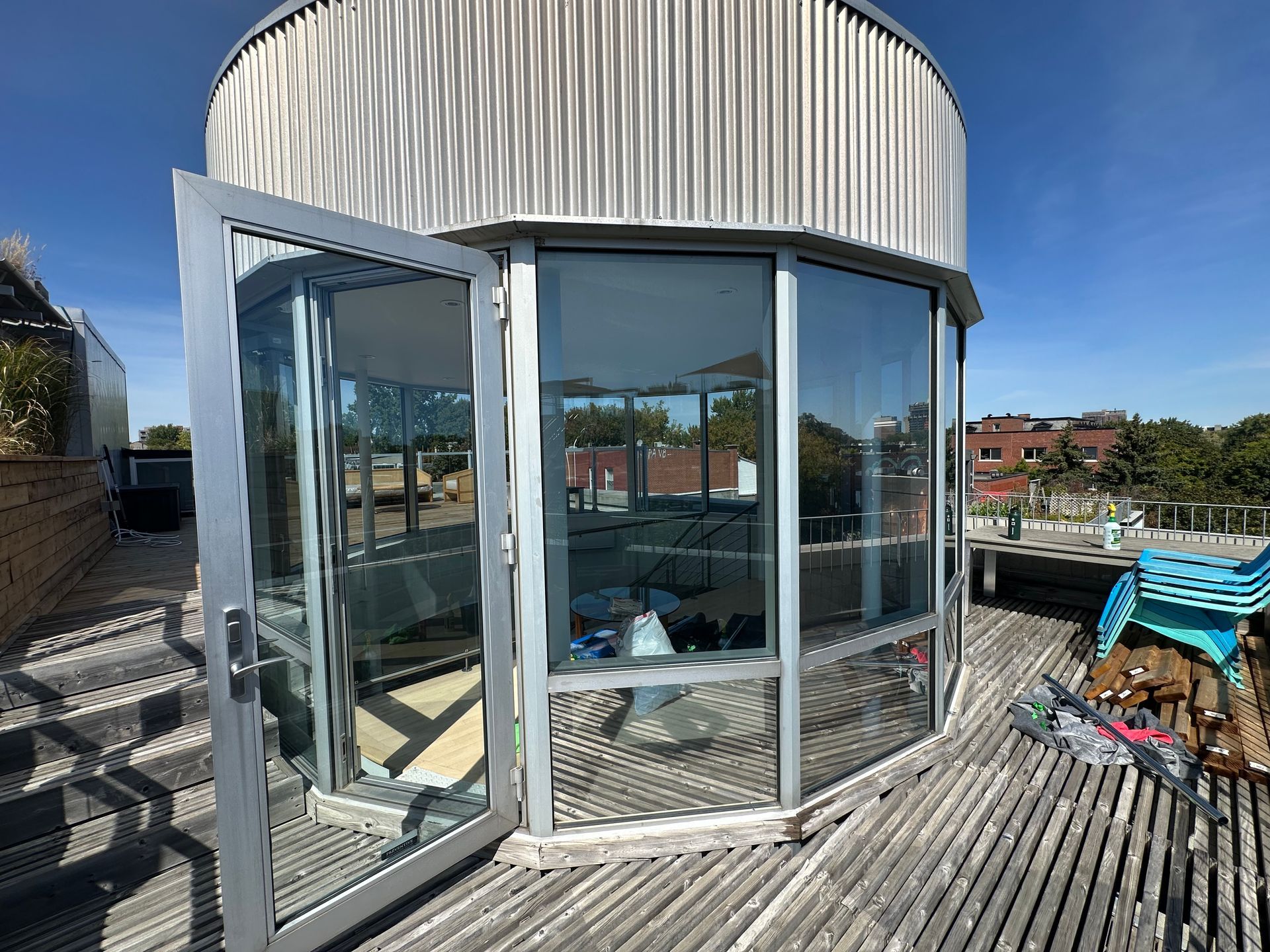 Une structure incurvée aux parois de verre sur un toit, avec des portes ouvertes, une terrasse en bois et un ciel bleu.
