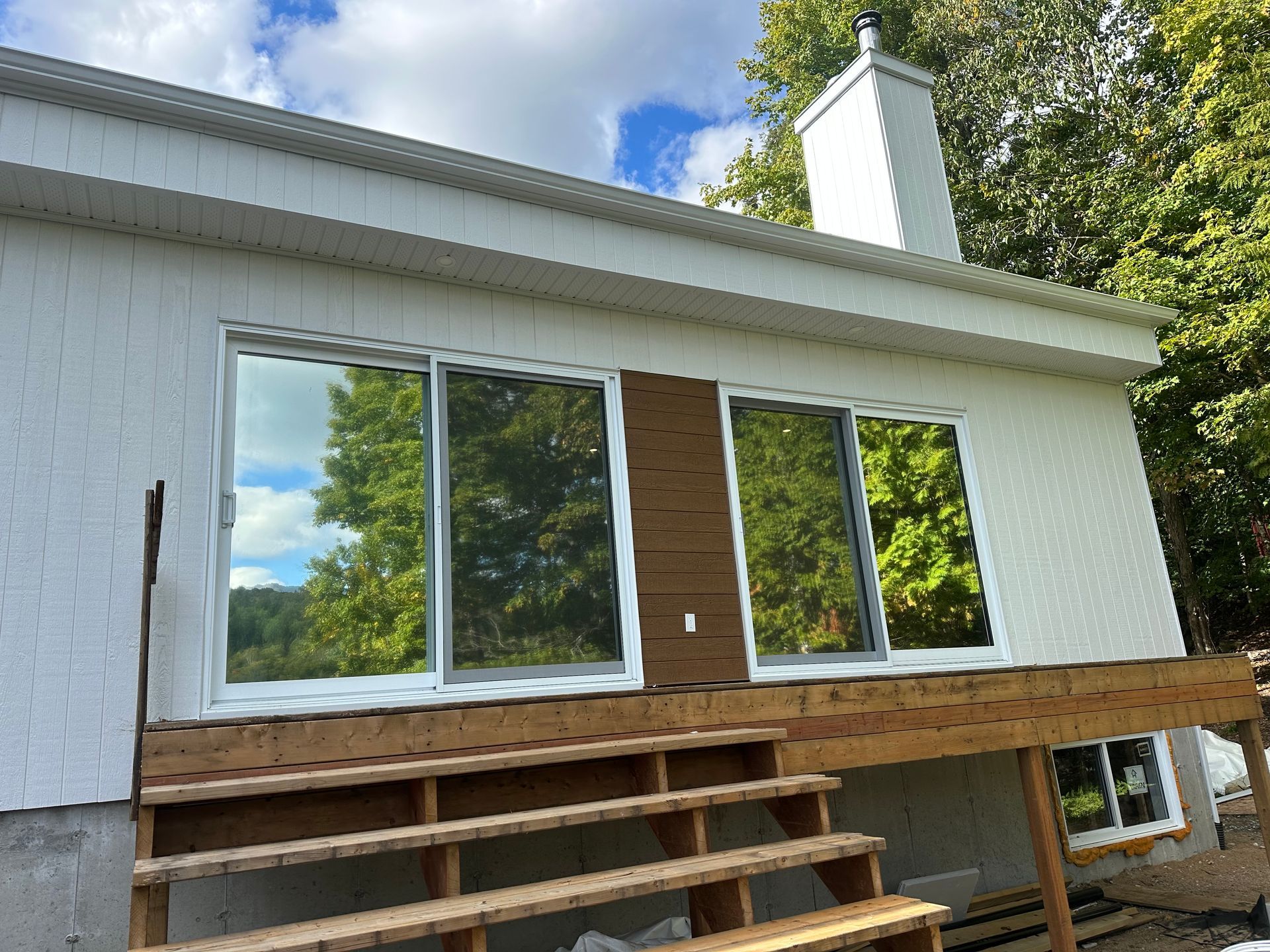 Extérieur d'une maison blanche avec de grandes portes coulissantes en verre, une terrasse en bois et une cheminée ; journée ensoleillée.