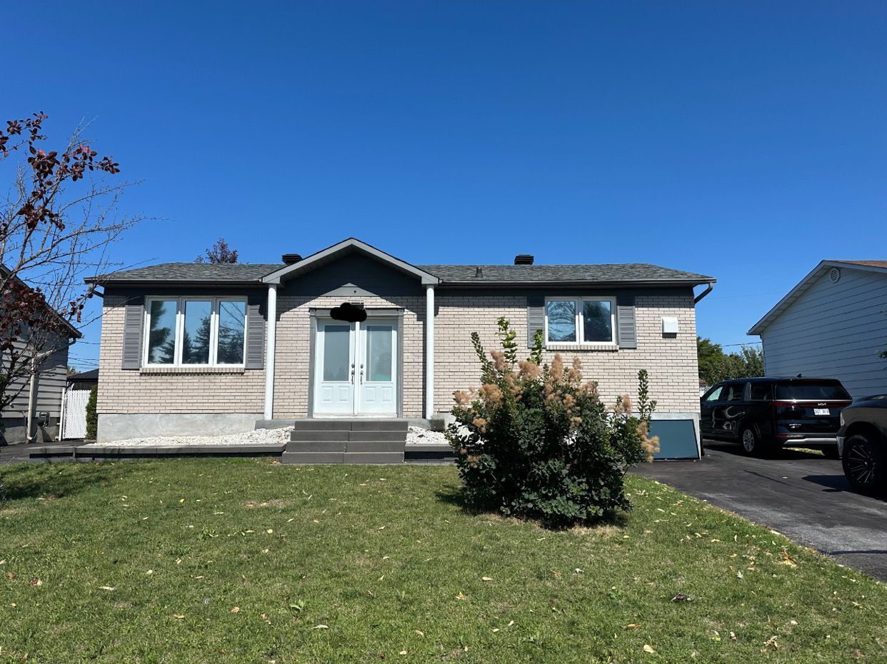 Maison à façade en briques grises, ciel bleu clair, pelouse et allée.