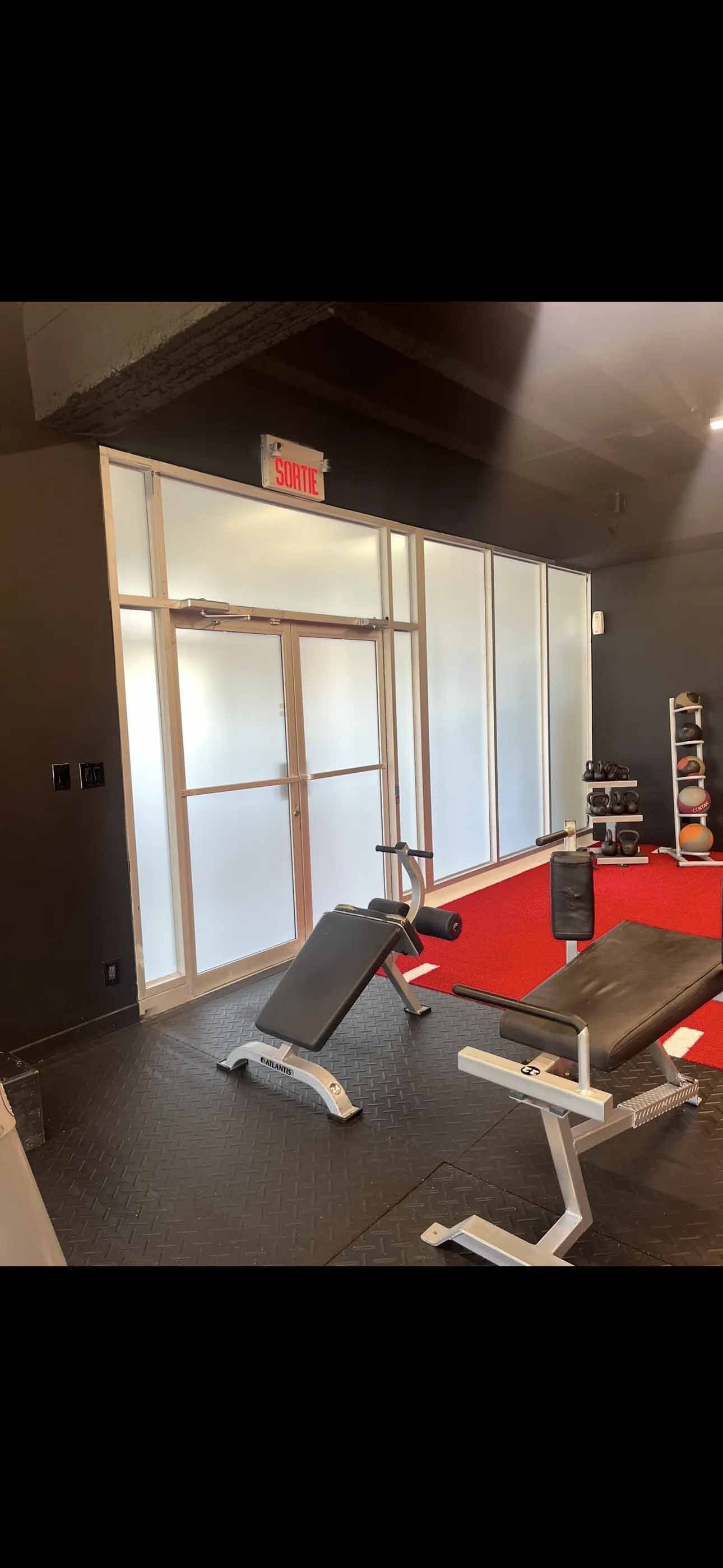 Intérieur d'une salle de sport avec deux bancs de musculation sur des tapis noirs. Mur en verre dépoli et sol rouge en arrière-plan.