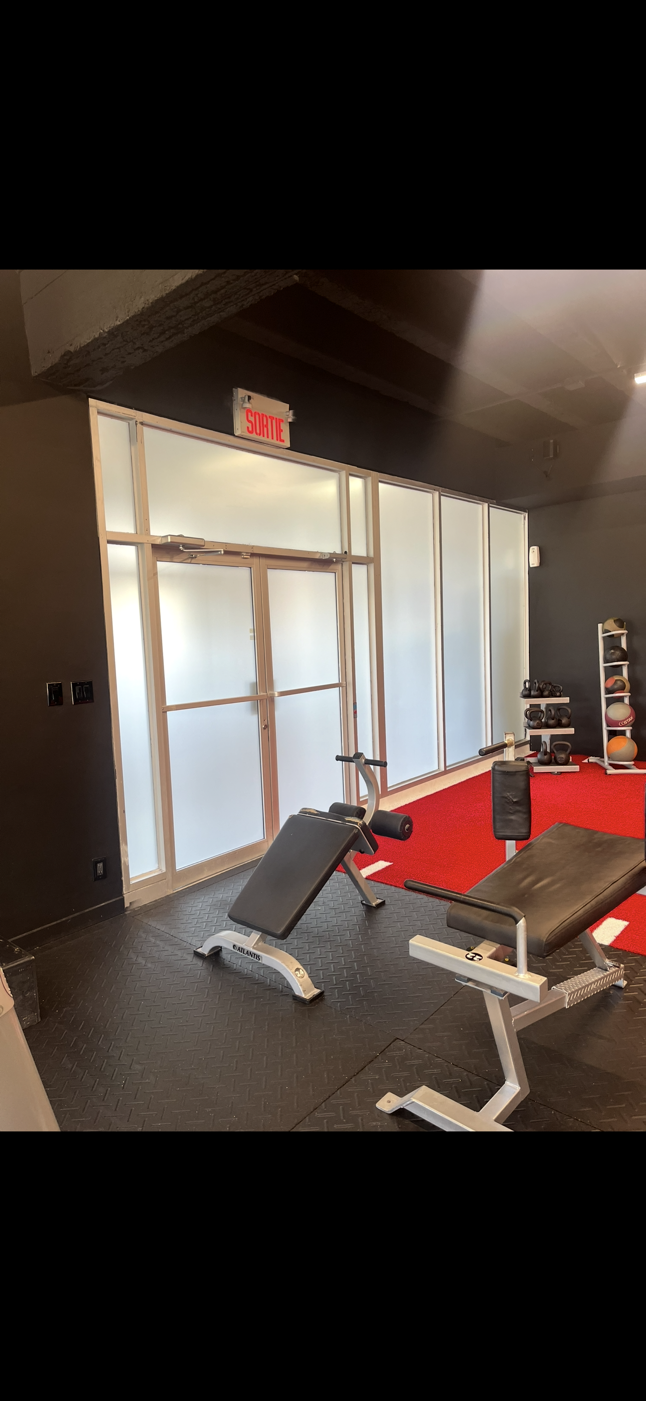 Intérieur d'une salle de sport avec deux bancs de musculation sur des tapis noirs. Mur en verre dépoli et sol rouge en arrière-plan.