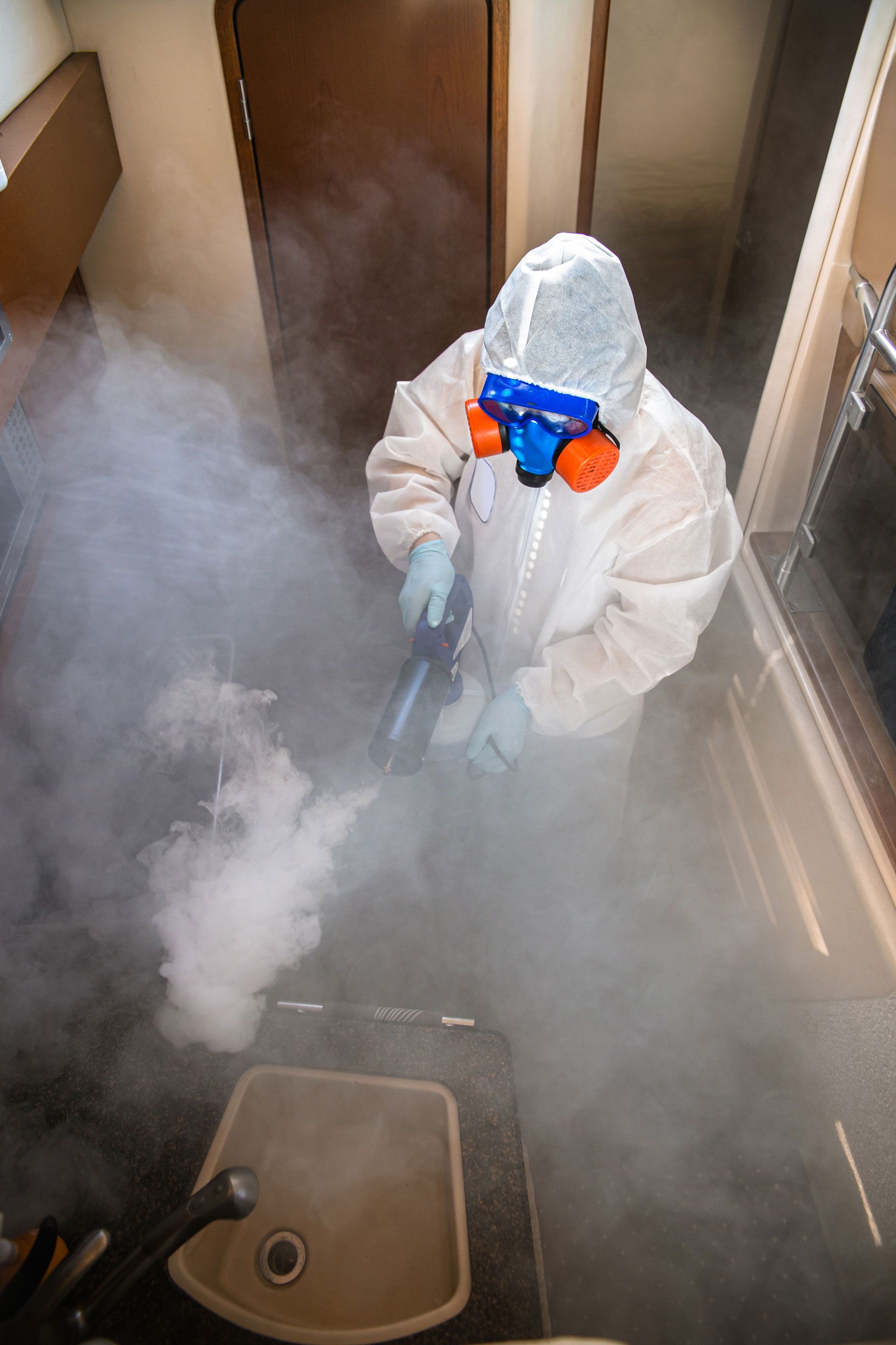 Person in protective suit spraying a room with mist; kitchen interior.