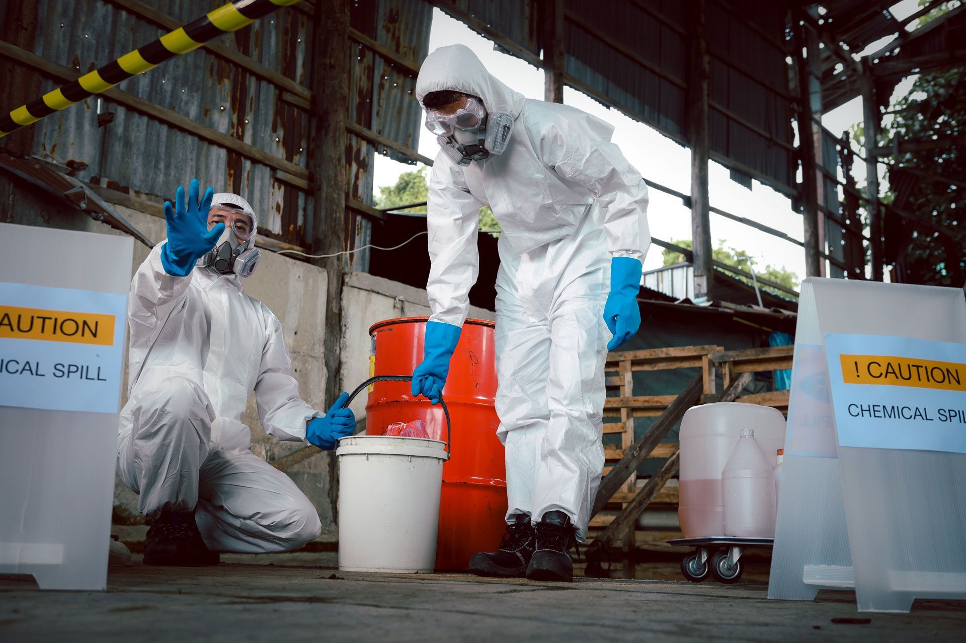 Two people in hazmat suits investigate a chemical spill near caution signs and a barrel.