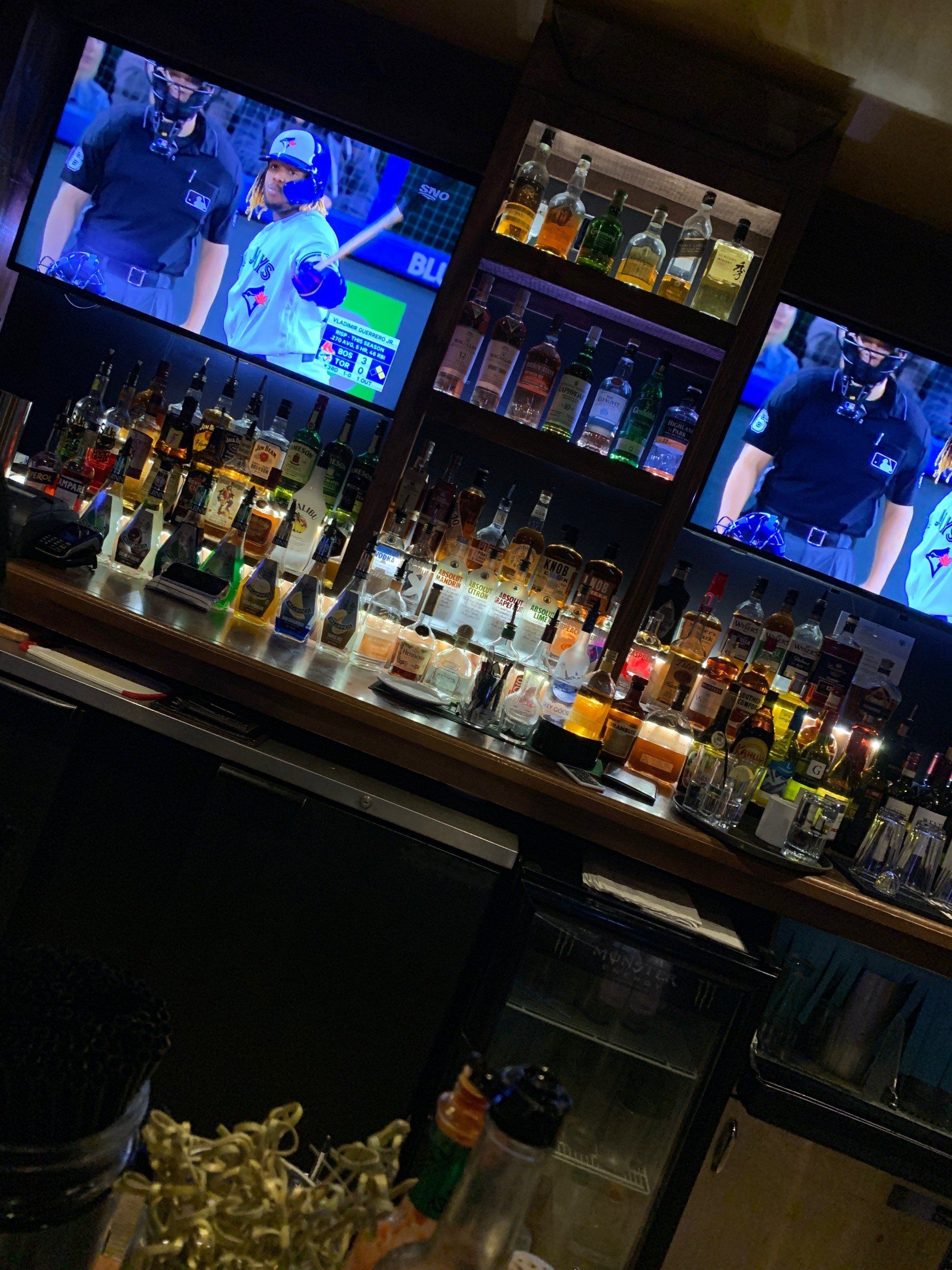 Bar interior with stocked shelves and two TVs showing a baseball game.