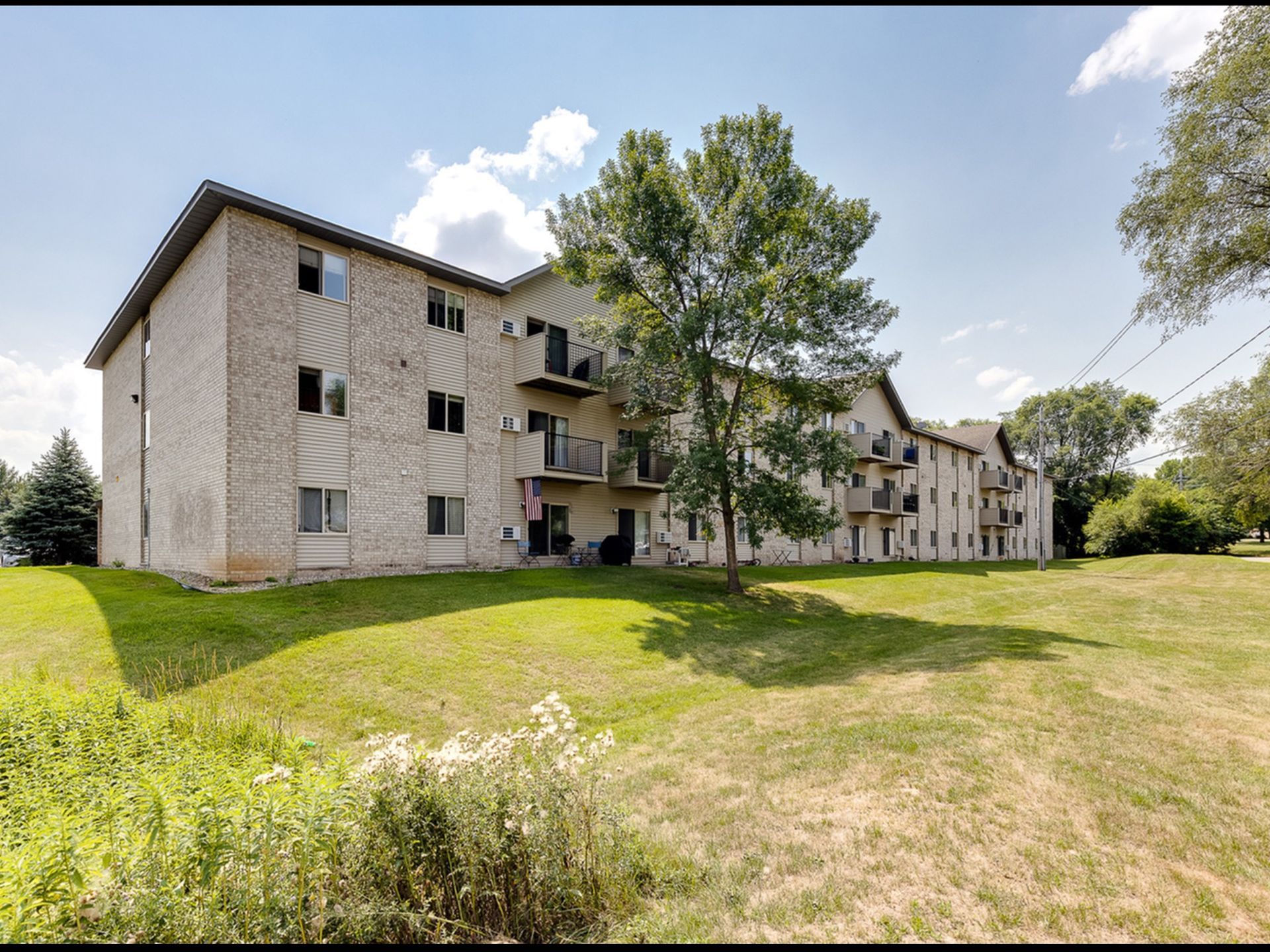 Exterior view of a beige brick apartment building with balconies and a grassy lawn.