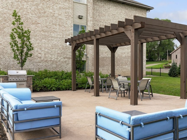 Outdoor community patio with blue cushioned seating under a wooden pergola near a brick building.