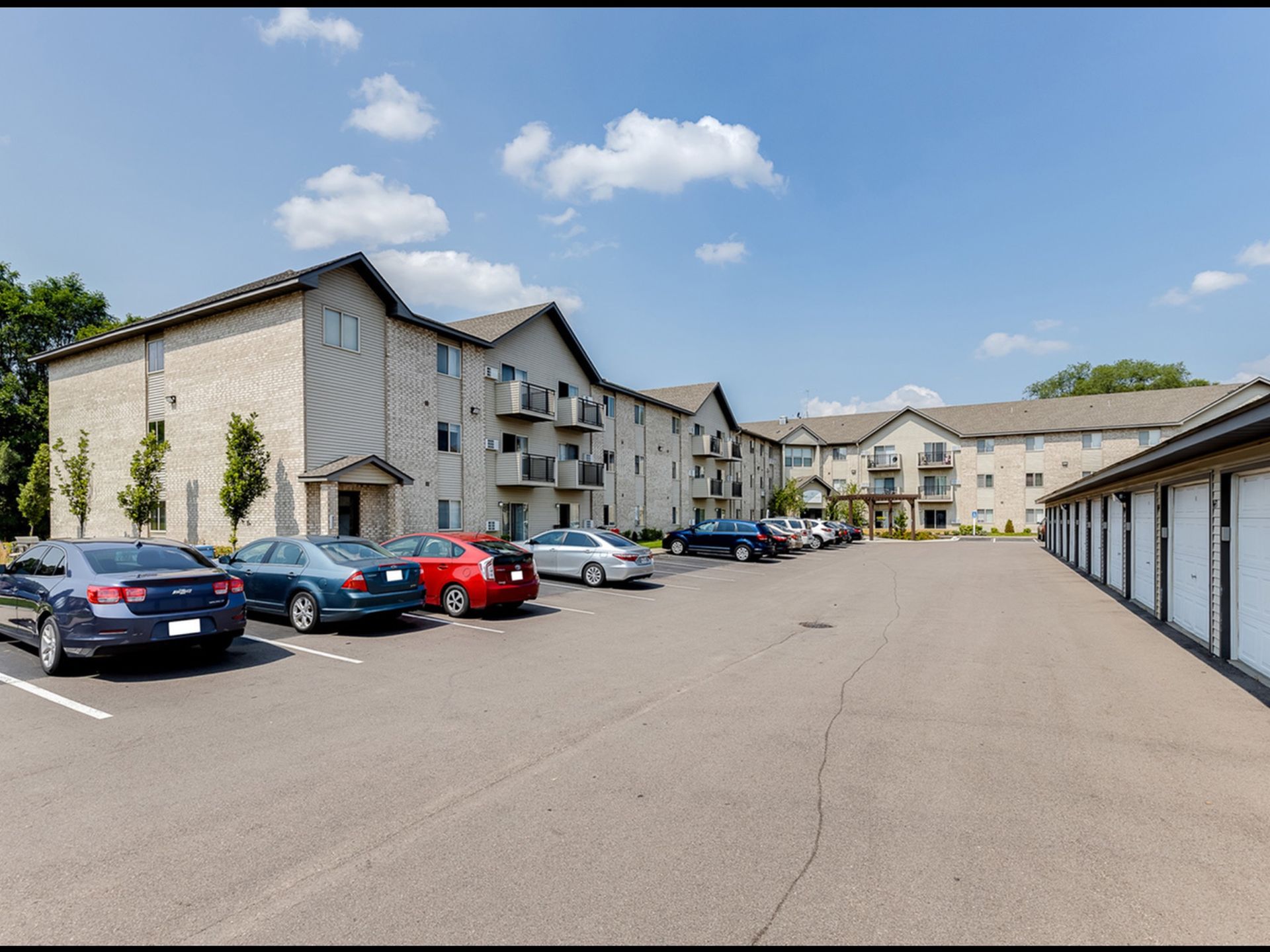Exterior view of a multi-building apartment community with a long parking lot and row of garages.