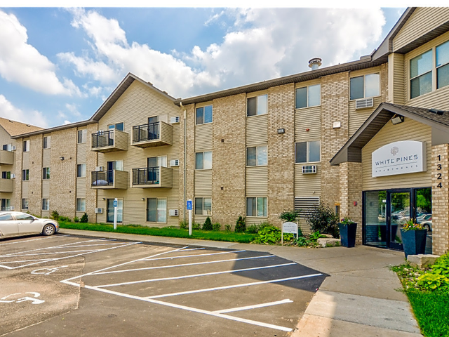 Exterior view of White Pines apartment building with beige brick, balconies, and a parking lot.