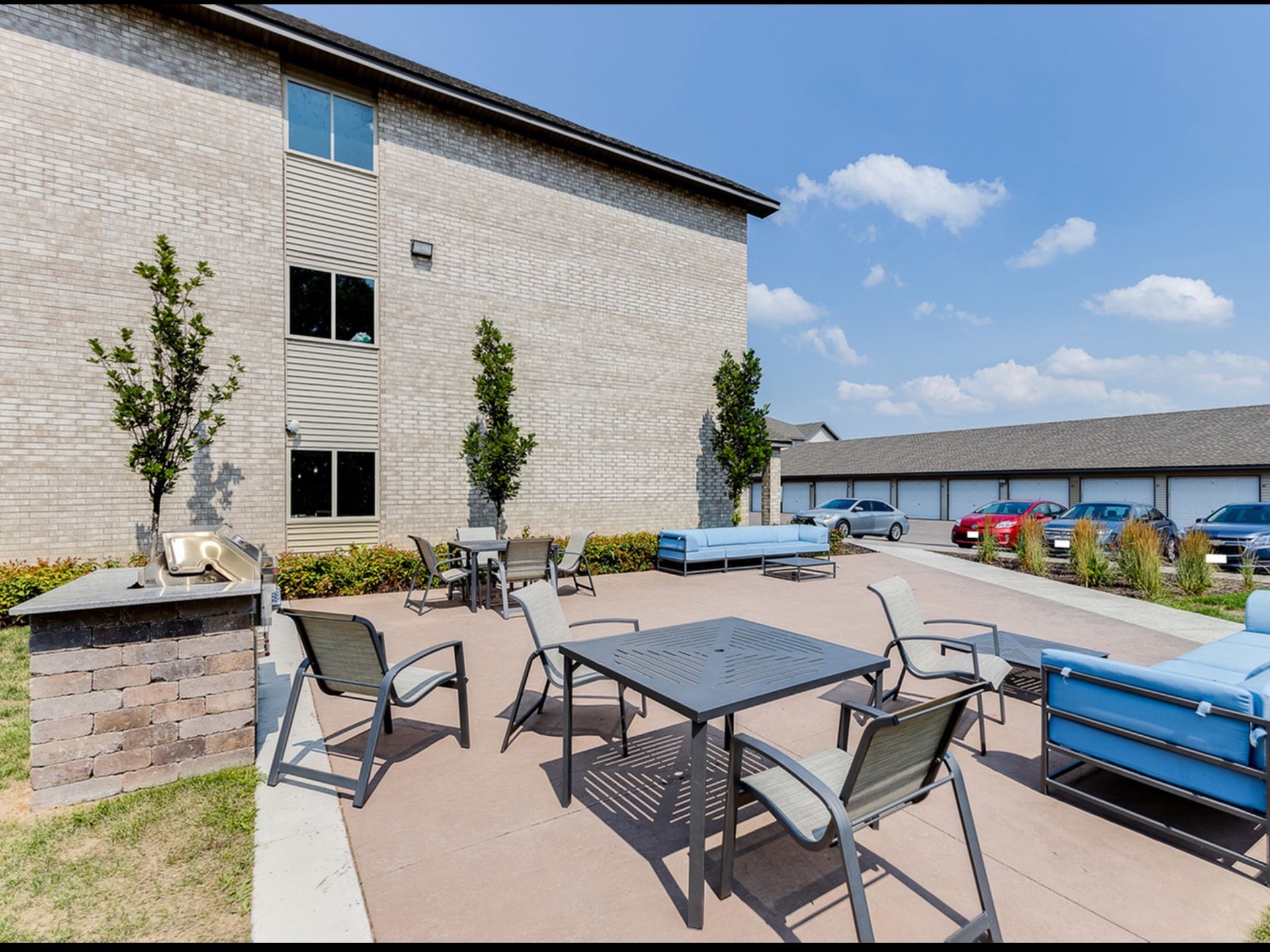 Outdoor resident courtyard with tables, chairs, a grill, and blue seating near the brick apartment building.