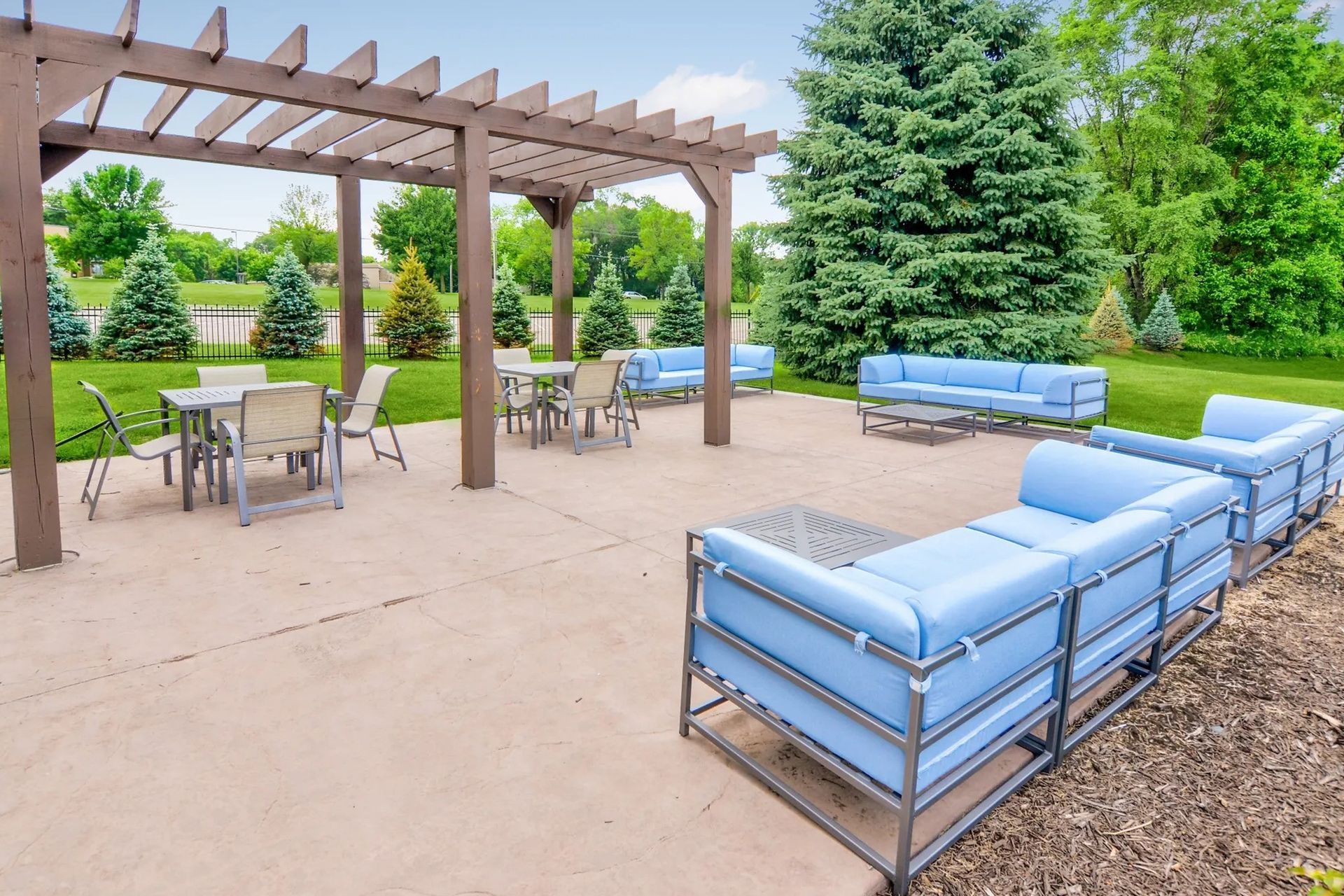 Patio with seating, pergola, and tables, surrounded by grass and trees.