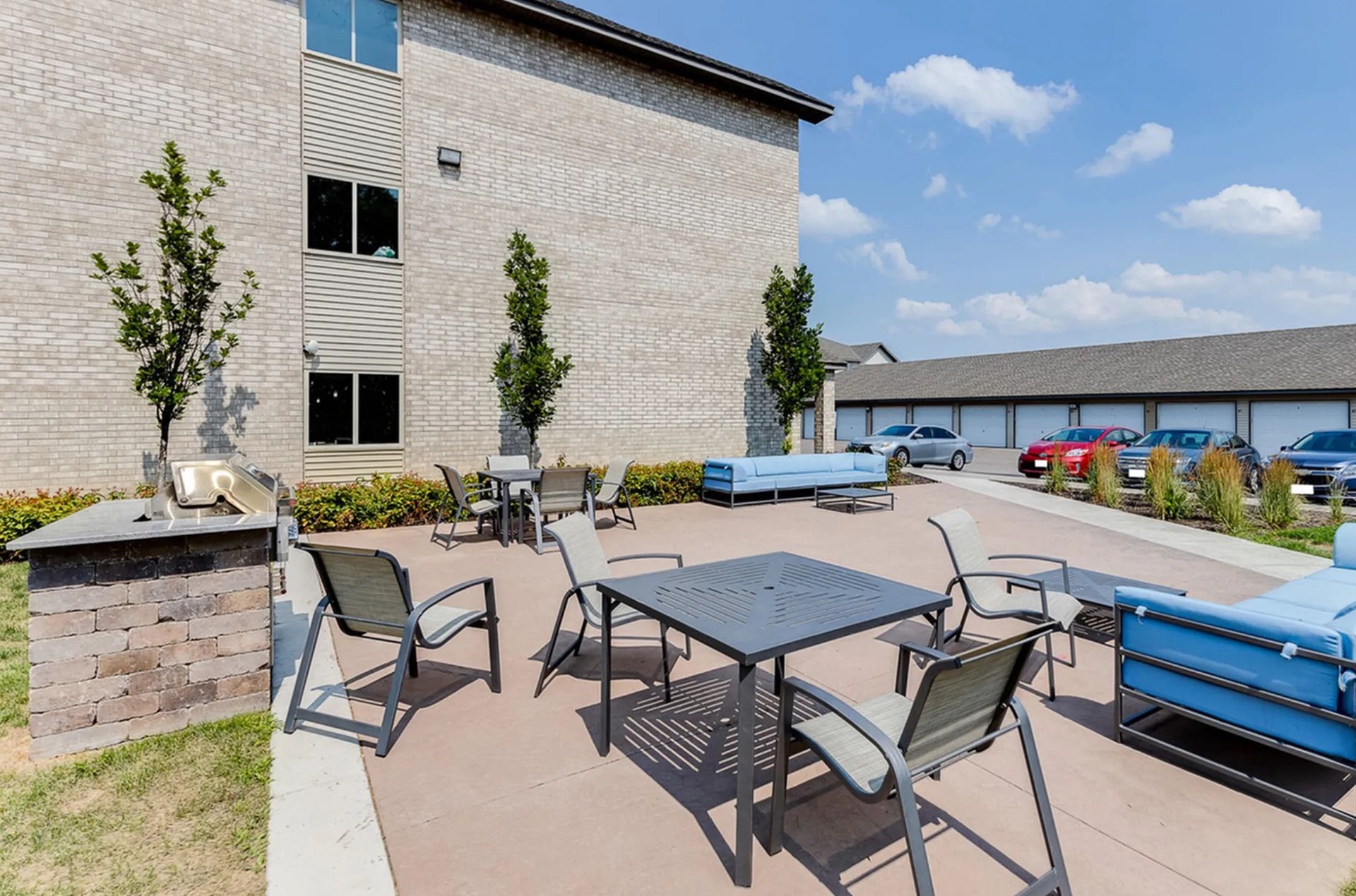 Outdoor patio with tables, chairs, and couches near a brick building and garage.