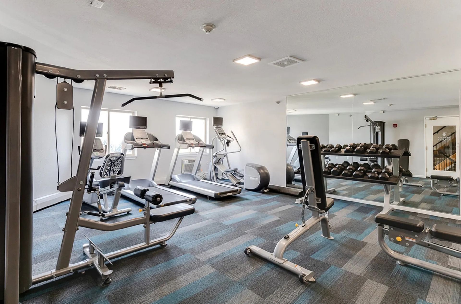 Gym with weight machines, treadmills, elliptical, and free weights, blue carpet.