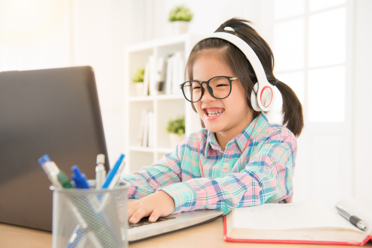 A little girl wearing headphones and glasses is using a laptop computer.