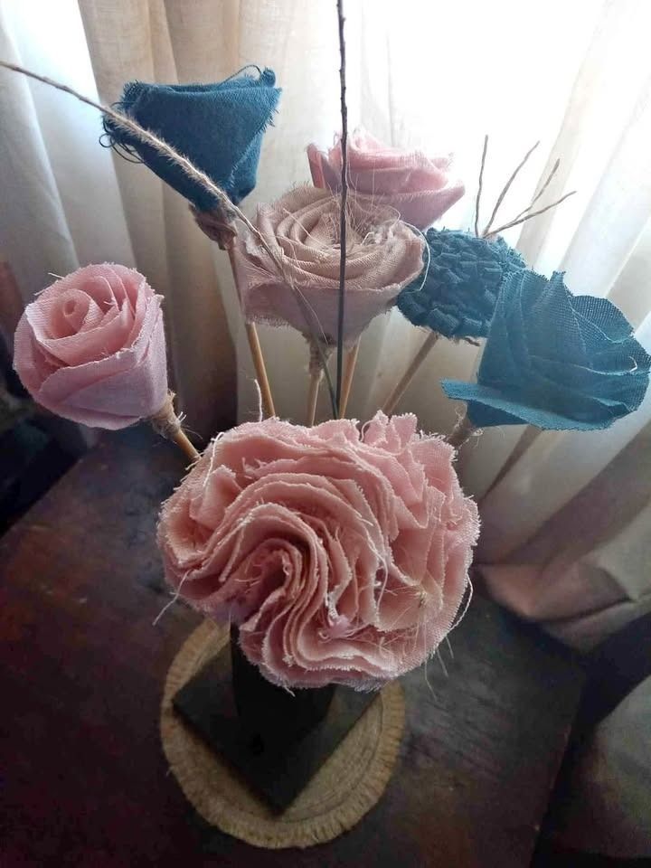 Flores de tela en tonos rosa y verde azulado, dispuestas en un jarrón sobre una mesa de madera.