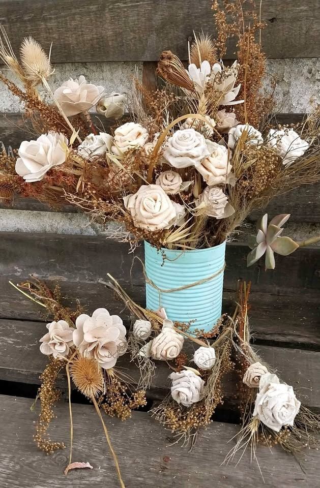 Arreglo de flores secas en una lata de color verde azulado sobre una superficie de madera, con ramos más pequeños al lado.
