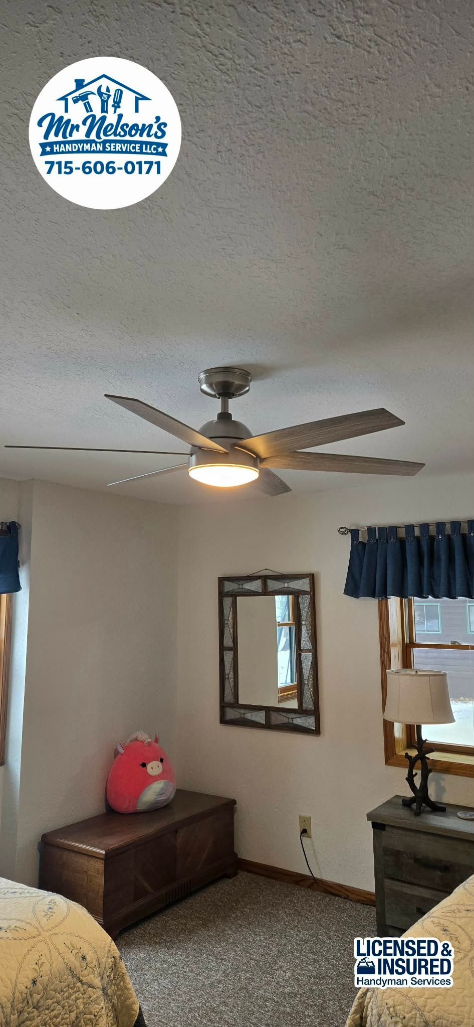 A bedroom with a lit ceiling fan, a small wooden trunk with a pink stuffed toy, a mirror, and a bedside lamp.