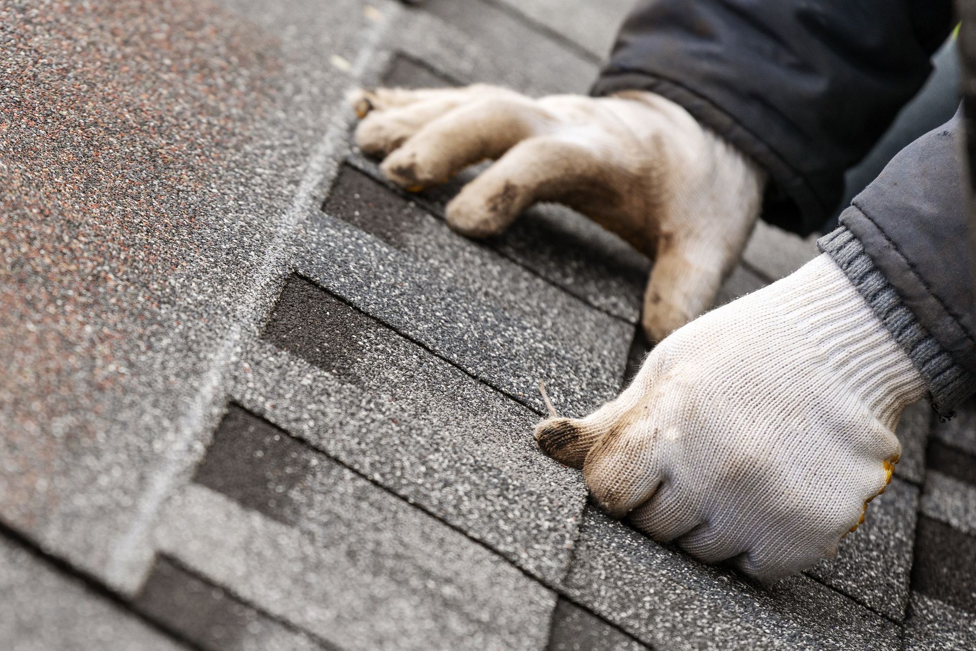 Gloved hands installing shingles on a rooftop with a hammer